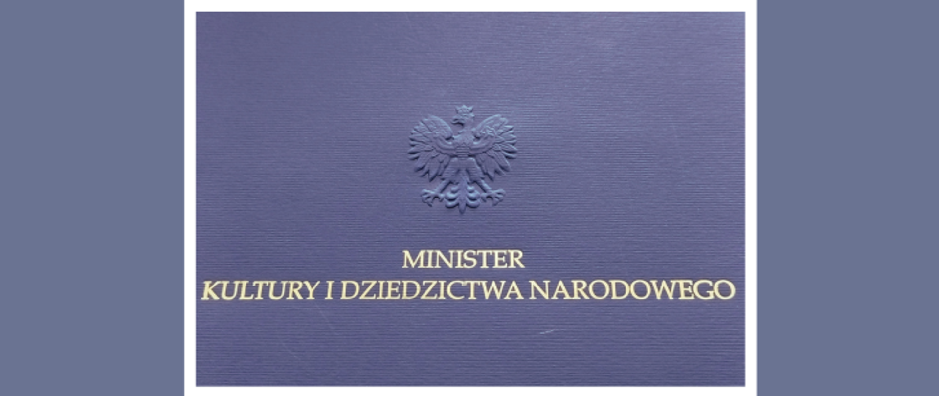 Obrazek pokazuje granatową okładkę ze złotym napisem i godłem Polski. Na środku górnej części okładki widać wypukłe, złocone godło Rzeczypospolitej Polskiej, czyli orła w koronie. Poniżej orła, wycentrowany, znajduje się złoty, drukowany napis w dwóch liniach: MINISTER w pierwszym rzędzie, a pod nim KULTURY I DZIEDZICTWA NARODOWEGO. Całość jest otoczona jasnoniebieską obwódką, co wygląda jak ramka. Okładka ma lekko fakturowaną strukturę. Złote elementy lśnią pod światło, a całość wygląda bardzo oficjalnie. Tło poza jasnoniebieską obwódką jest ciemnofioletowe.
