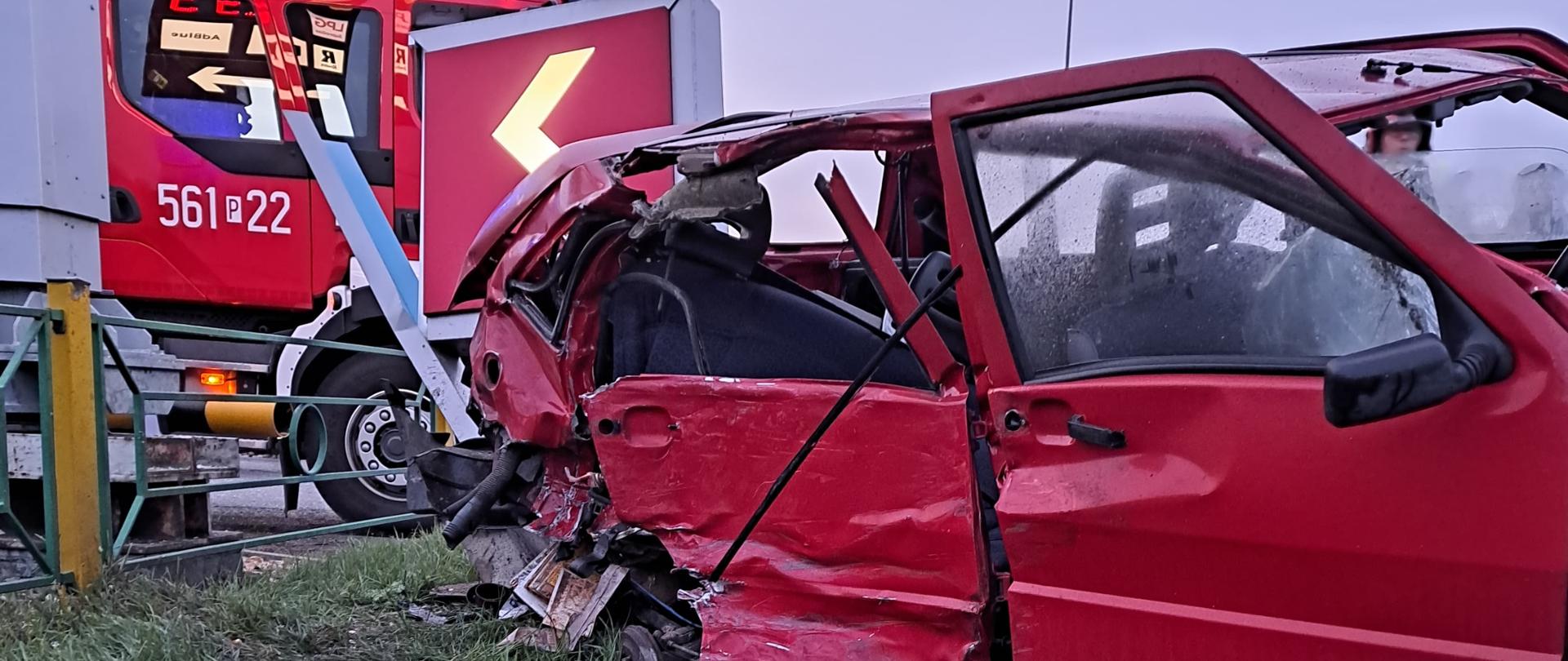 Czerwony samochód osoby ze zniszczonym bokiem stoi na uszkodzonym mostku przejazdowym za nim widoczny wóz strażacki