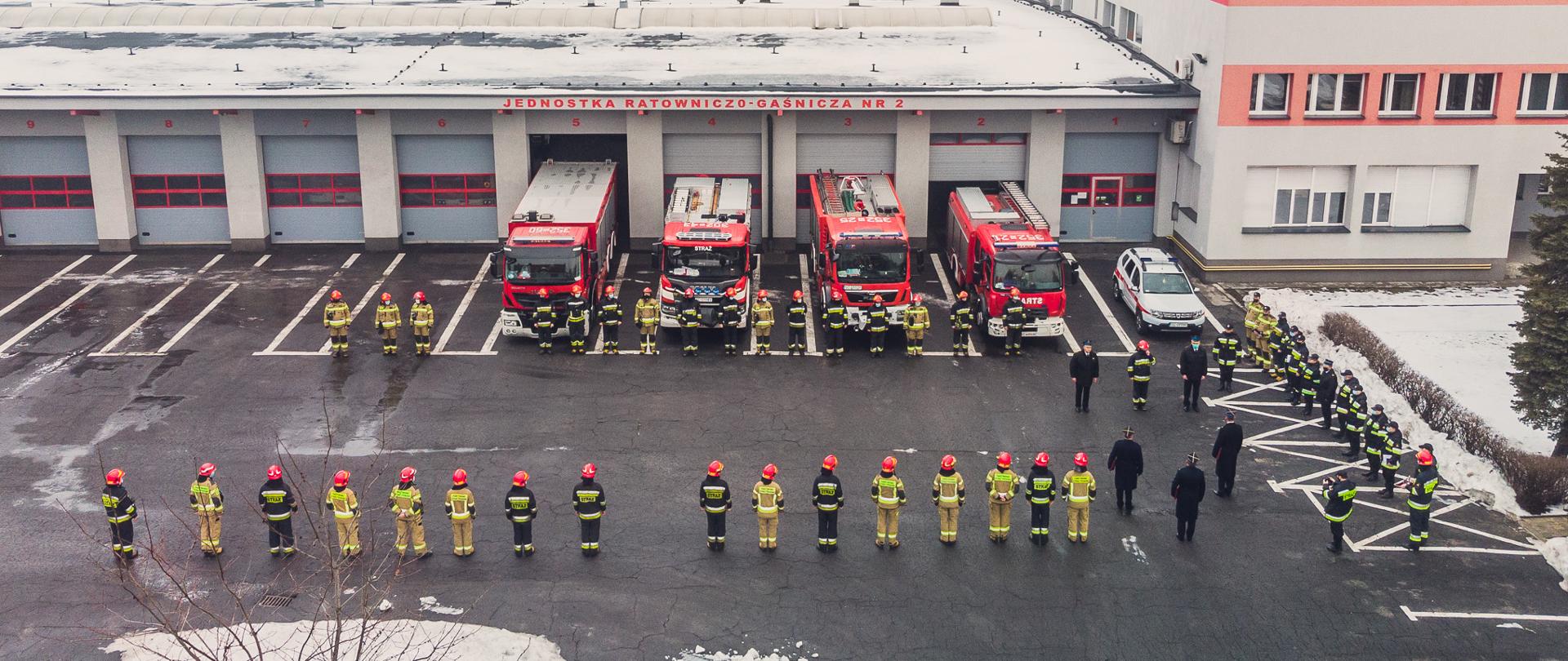 Zdjęcie przedstawia funkcjonariuszy Państwowej Straży Pożarnej podczas uroczystej zmiany służby. Na zdjęciu widoczni są funkcjonariusze Państwowej Straży Pożarnej z lotu ptaka. Strażacy stoją w dwóch szeregach przodem do siebie na tle budynku Komendy Miejskiej Państwowej Straży Pożarnej w Częstochowie. Przed budynek wyprowadzonych jest 5 pojazdów pożarniczych