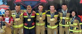 Zdjęcie 1: Grupa siedmiorga polskich strażaków i zawodników w ubraniach specjalnych pozuje z medalami przed grafiką „Firefighter Challenge Championship Series”.