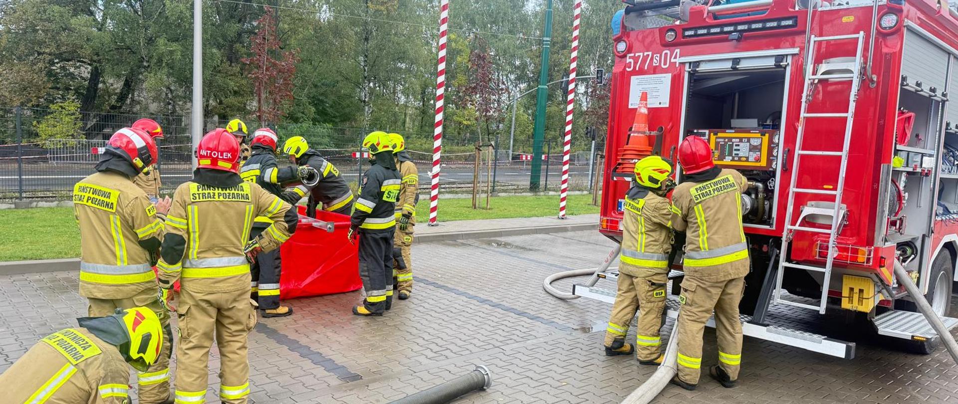 Grupa strażaków prowadzi ćwiczenia przy wozie strażackim, podłączając węże z wykorzystaniem przenośnego zbiornika na wodę.