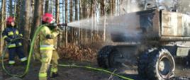 Na zdjęciu dwóch strażaków. Jeden w piaskowym a drugi ciemnym granatowym ubraniu. Strażacy stoją przy spalonej koparce. Jeden ze strażaków dogasza koparkę