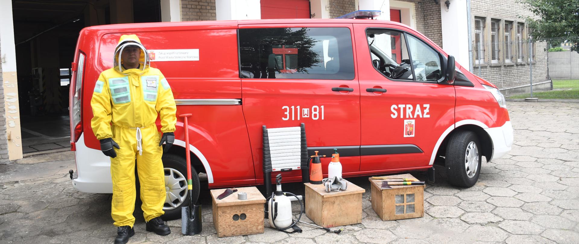 Na zdjęciu widać strażaka w stroju ochronnym koloru żółtego oraz typowy sprzet do usuwania gniazd pszczół, drabinka, siekiera, maska ochronna, szpadel. W tle samochód straży pożarnej typu bus.