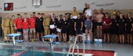 Grupa kilkudziesięciu osób stoi ustawiona w kilku rzędach na pływalni, pozując do wspólnego zdjęcia. Po lewej stronie znajdują się osoby w beżowych mundurach służbowych, obok nich grupa w czerwonych koszulkach. Centralnie i po prawej stronie stoją drużyny w czarnych strojach sportowych oraz kilka osób w strojach kąpielowych trzymających puchary i dyplomy, w tym część ustawiona na podium. Na pierwszym planie widać krawędź basenu z blokami startowymi. W tle znajdują się zegary, tablice informacyjne i ściana pływalni.
