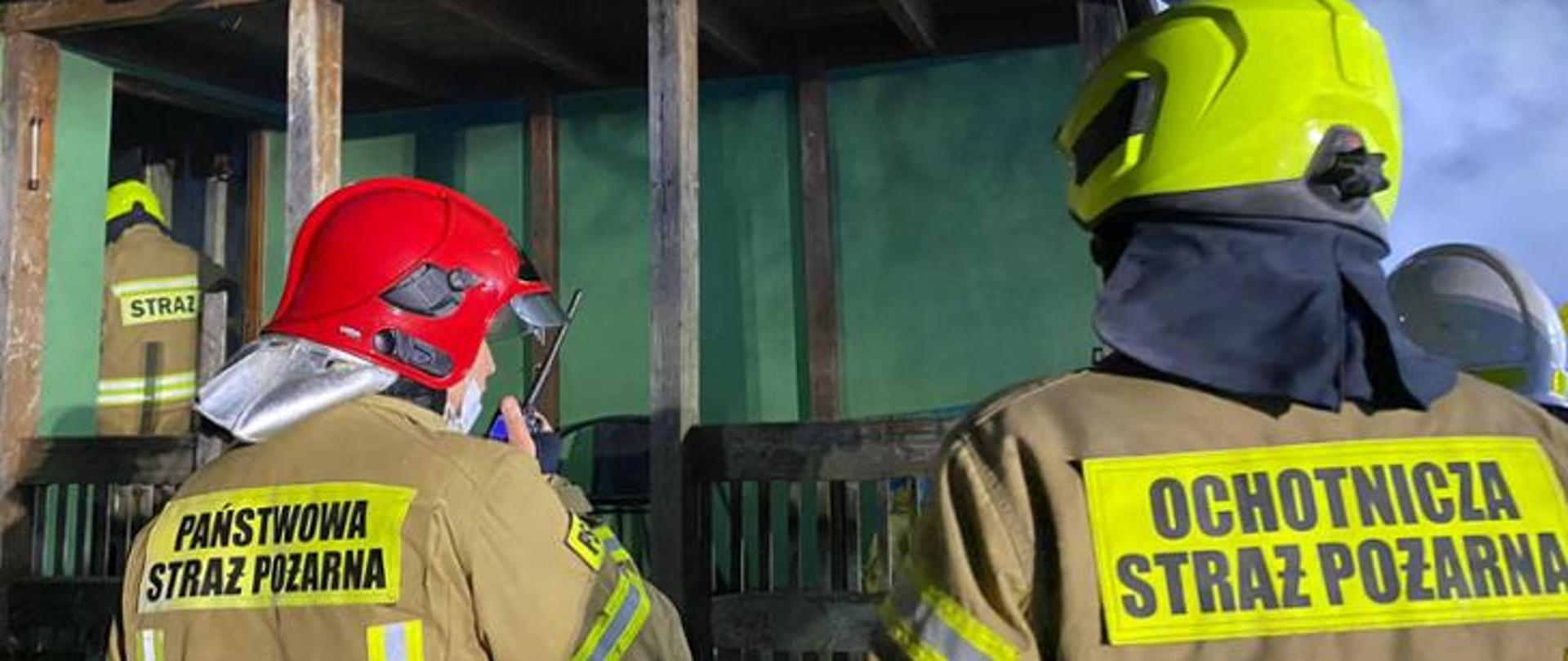 Dwóch strażaków odwróconych tyłem, ubranych w ubrania specjalne, na plecach napisy Państwowa Straż Pożarna oraz Ochotnicza Straż Pożarna