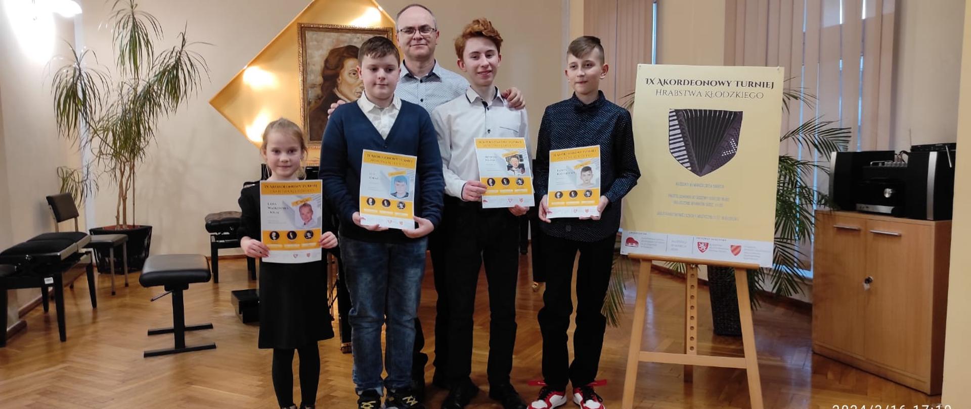 Zdjęcie przedstawia czterech laureatów IX Akordeonowego Turnieju Hrabstwa Kłodzkiego oraz ich nauczyciela. Dzieci trzymają kolorowe dyplomy laureata na tle portretu Chopina i akcesoriów muzycznych oraz plakatu z informacją o turnieju. 