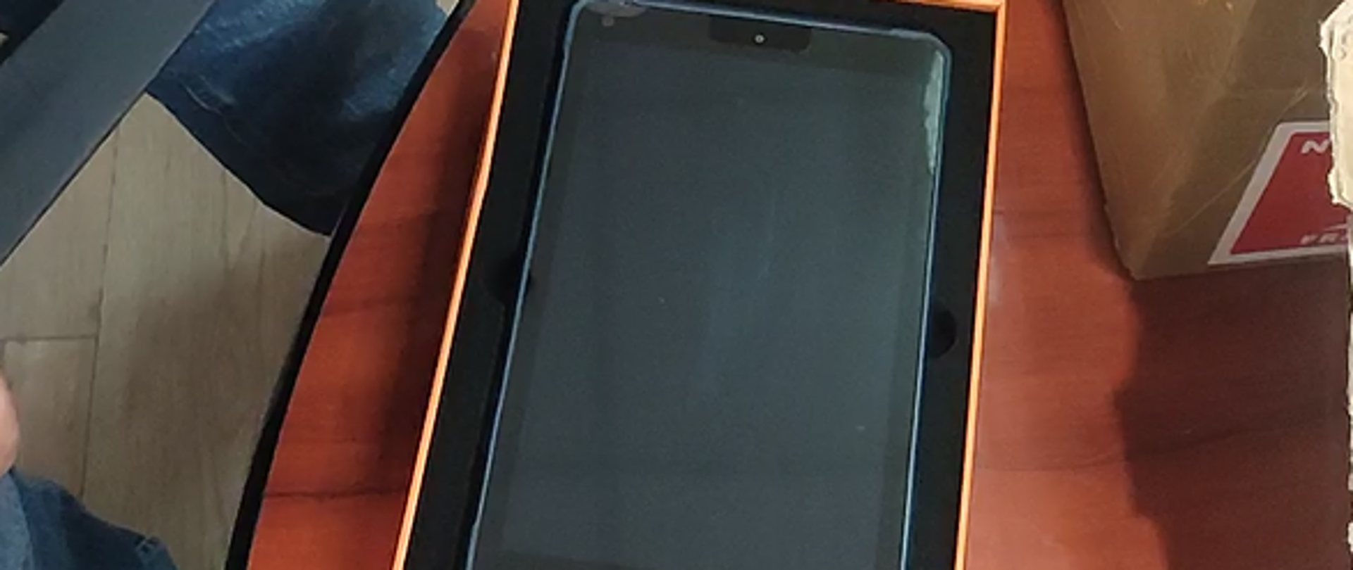 Ręka osoby wyjmującej tablet z czarno-pomarańczowego pudełka, obok otwartego kartonu z folią bąbelkową na drewnianym stole.