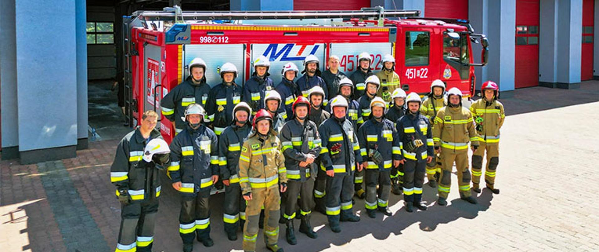 24 ratowników ochotniczych straży pożarnych ubranych w ubrania typu moratex mają na głowie hełm pozuje do pamiątkowego zdjęcia na tle budynku garażowego komendy powiatowej PSP w Łowiczu w tle za nimi stoi samochód ratowniczy barwy czerwonej z drabiną na dachu 