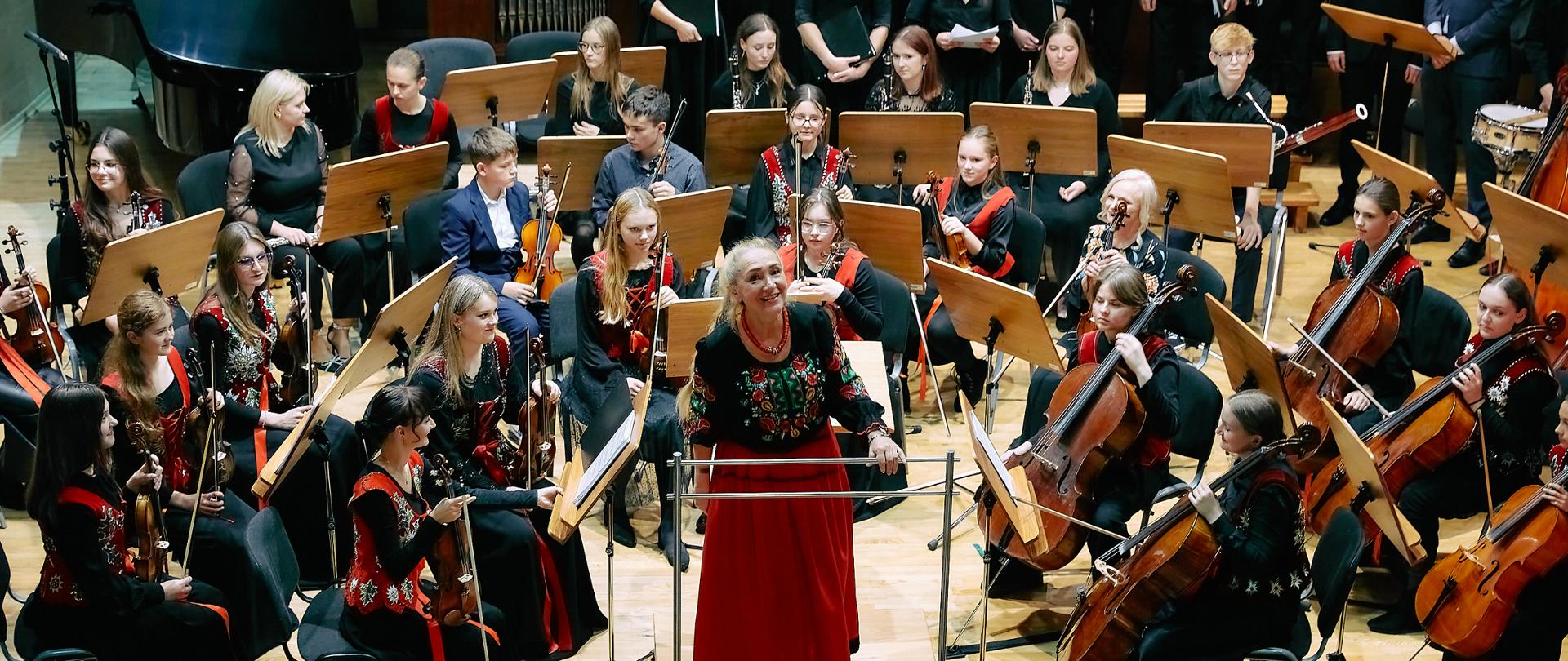 Zdjęcie zrobione z balkonu, obejmujące całą scenę sali koncertowej. Na scenie orkiestra ubrana w stroje regionalne, na środku na podeście skierowana do publiczności uśmiechnięta pani dyrygent. Za orkiestrą w prawej części sali stoi chór towarzyszący orkiestrze, z lewej strony fortepian a przy nim pianistka. W tle organy.