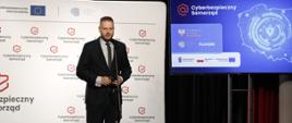 Minister cyfryzacji Janusz Cieszyński stoi na scenie i przemawia do mikrofonu, za jego plecami baner z multiplikacją „Cyberbezpieczny Samorząd”