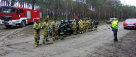 Zdjęcie przedstawia strażaków w lesie podczas ćwiczeń. Instruktor omawia techniki usuwania skutków silnych wiatrów i nawałnic. Na zdjęciu widać również samochody strażackie. 