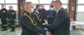 Zdjęcie przedstawia strażaków ubranych w granatowe mundury wyjściowe podczas wręczania odznaczeń i uścisków dłoni na uroczystej zbiórce w pomieszczeniu garażowym.