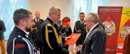 Pomorski komendant wojewódzki Państwowej Straży Pożarnej trzyma czerwoną teczkę przed nim stoi mężczyzna obok strażacy Państwowej Straży Pożarnej ochotniczych straży pożarnych oraz zaproszeni goście.