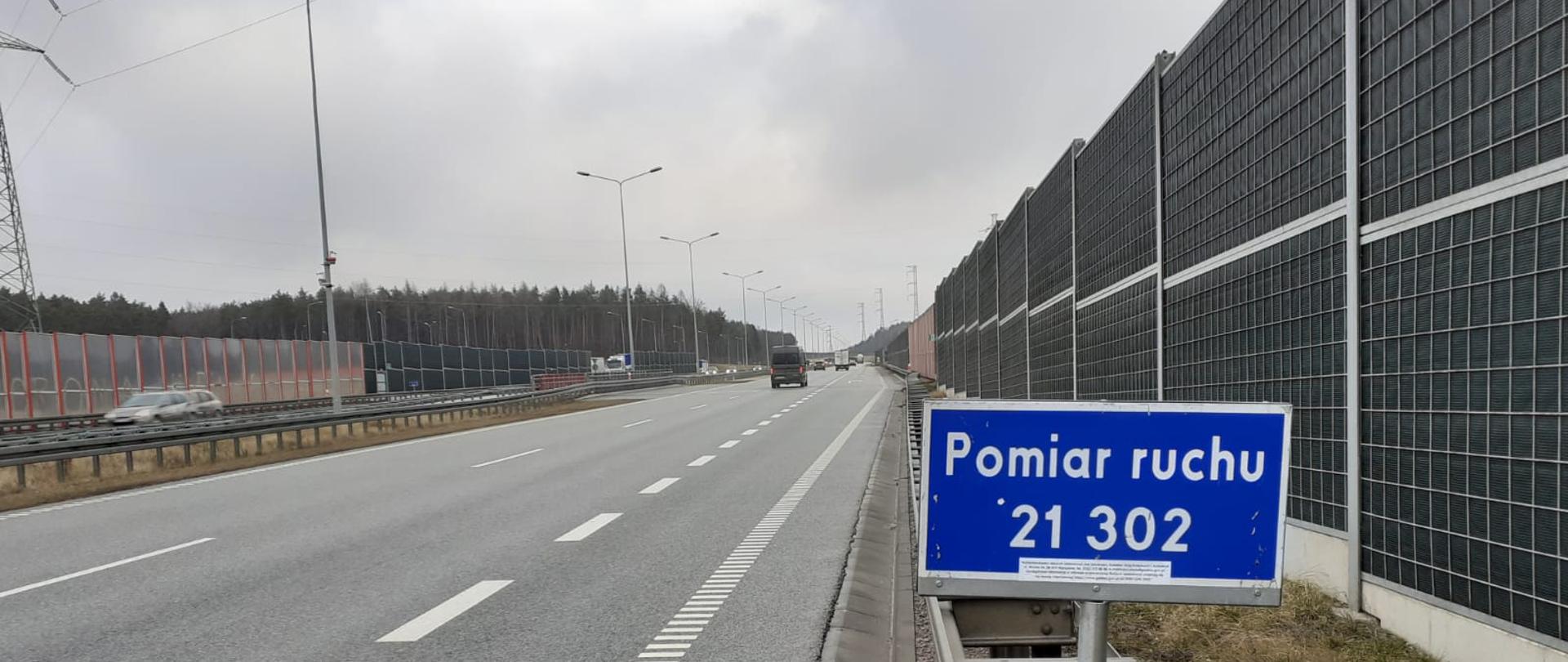 Tabliczka informująca o Generalnym Pomiarze Ruchu przy dwujezdniowej drodze. Przy jezdniach bariery i ekrany akustyczne. 