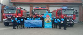 Zdjęcie przedstawia czternastu funkcjonariuszy Komendy Powiatowej Państwowej Straży Pożarnej w Gołdapi wyposażonych w niebieskie akcenty (balony, banery) na tle pojazdów pożarniczych z włączonymi niebieskimi sygnałami świetlnymi, solidarnych z osobami ze spektrum autyzmu
