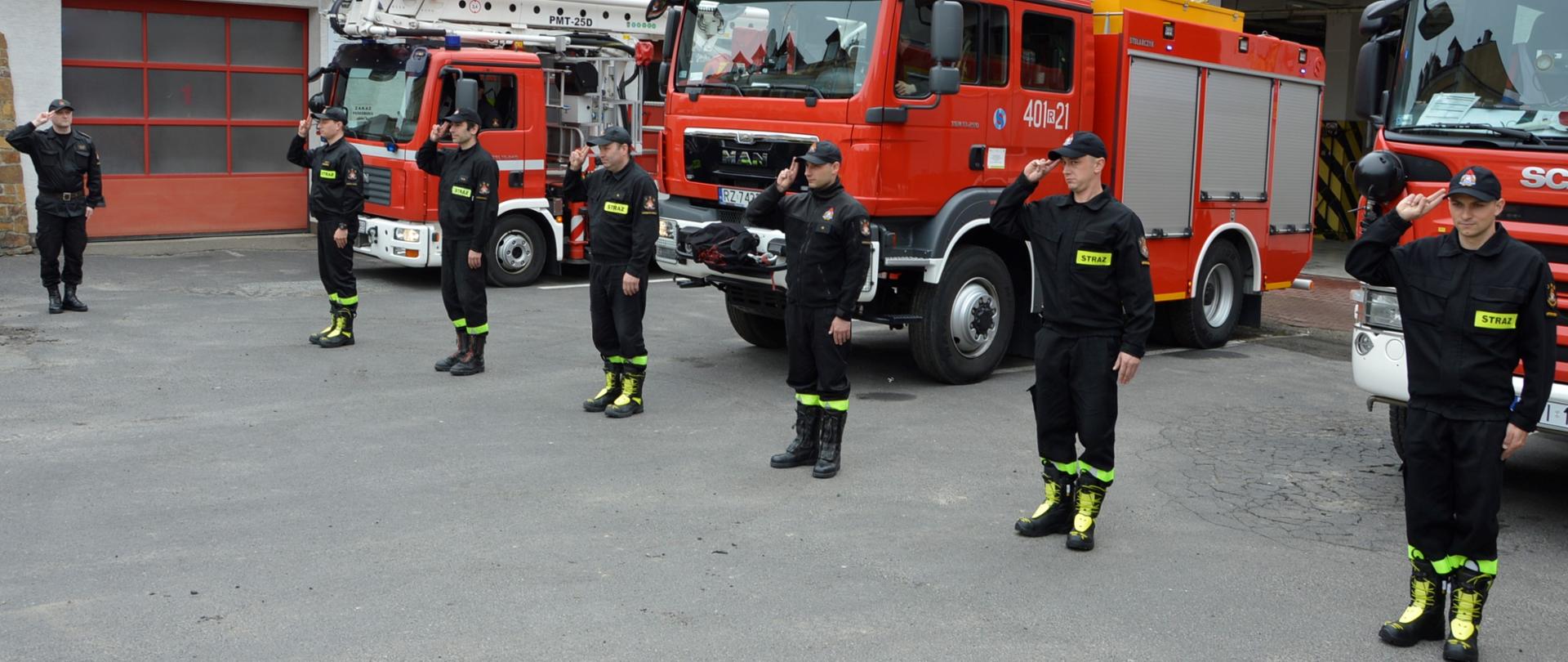 Na zdjęciu widać funkcjonariuszy KP PSP w Ustrzykach Dolnych którzy stoją przed komendą oddając honory. Za nimi widać samochody pożarnicze z włączonymi sygnałami świetlnymi a w tle budynek komendy.