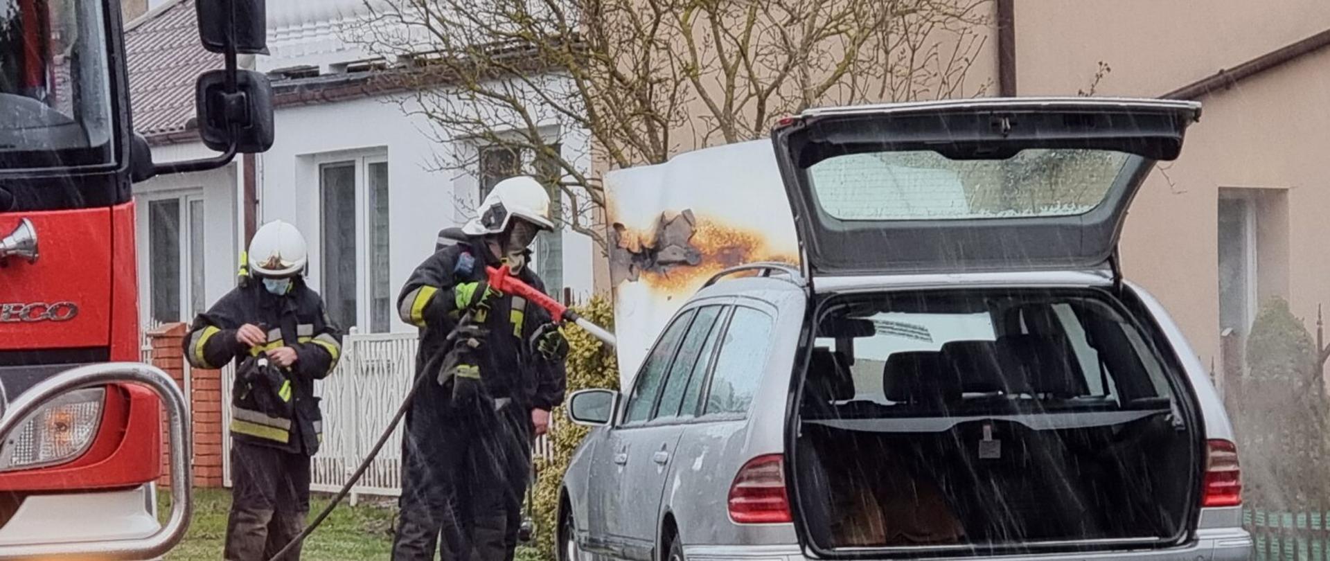 Zdjęcie przedstawia akcję gaśniczą w której dwóch strażaków gasi pojazd marki mercedes stojący na poboczu.W tle widać zabudowania.