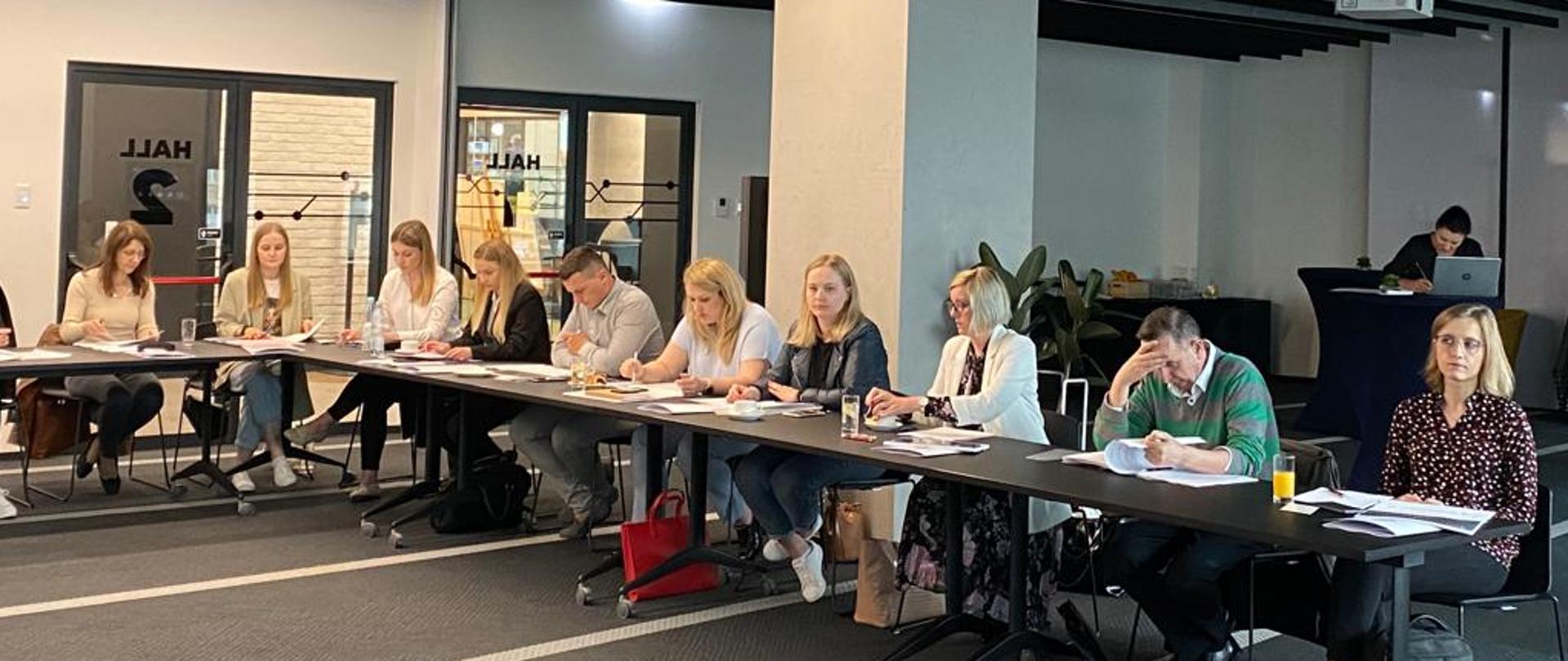 Druga część uczestników warsztatów siedzący przy stołach i analizujących materiały szkoleniowe. W tle kącik kawowy.