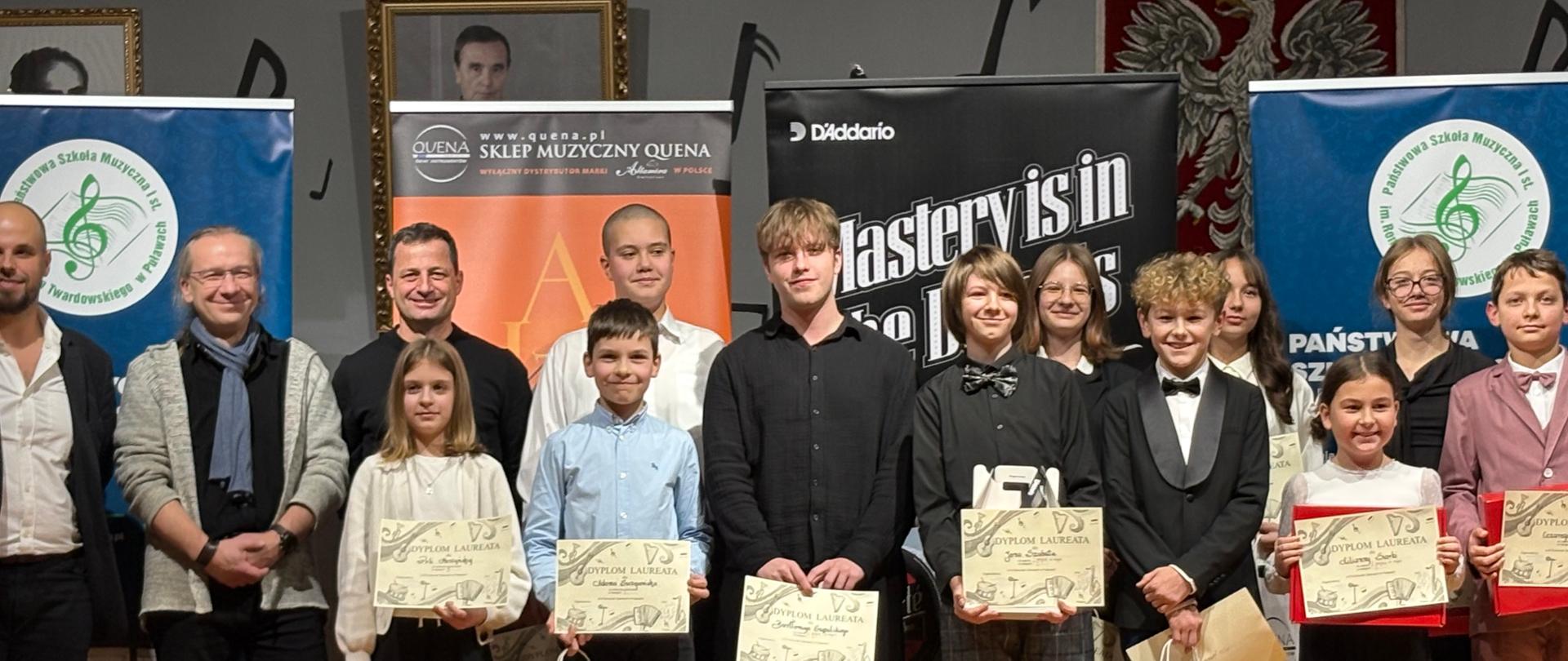 Uczniowie naszej szkoły Bartłomiej Szypulski i Jan Szabat stoją na scenie z nagrodami i dyplomami za zdobycie I miejsca exequo.
Na zdjęciu widać laureatów kategorii 1. Na scenie stoi kilkanaście osób w różnym wieku – dzieci, młodzież i dorośli - członkowie jury, dyrektor szkoły, sponsor. Kilka osób trzyma dyplomy i torby z nagrodami. W tle są banery sponsorów i sklepu muzycznego. Drewniana scena.
