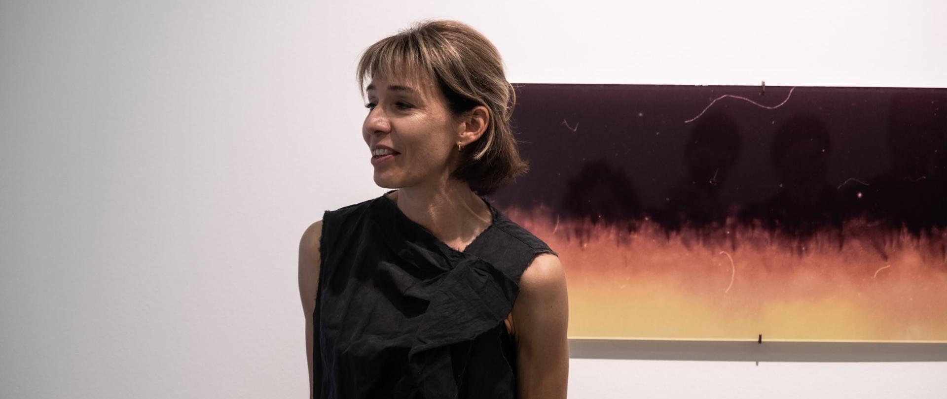 Poljska fotografinja Katerina Kouzmitcheva stoji v galerijskem prostoru pred eno od razstavljenih fotografij. Z nasmehom se obrača proti obiskovalcem odprtja razstave in predstavlja svoje delo. Za njo je vidna fotografija iz cikla razstave »Oprosti, ker se nisem oglasila«.
