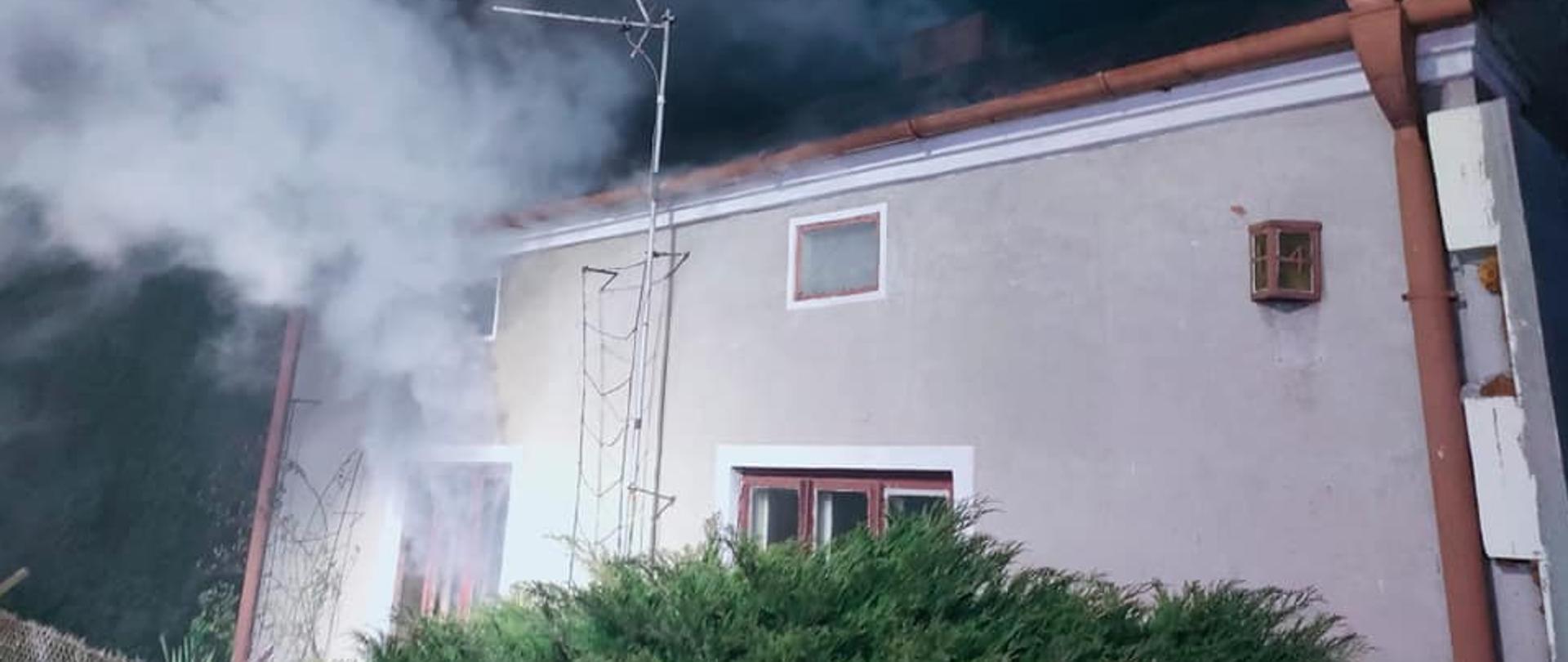 Na zdjęciu widać parterowy murowany budynek mieszkalny. Z okien oraz komina wydobywa się dym. Przed budynkiem widać zielony krzew. Jest ciemno.