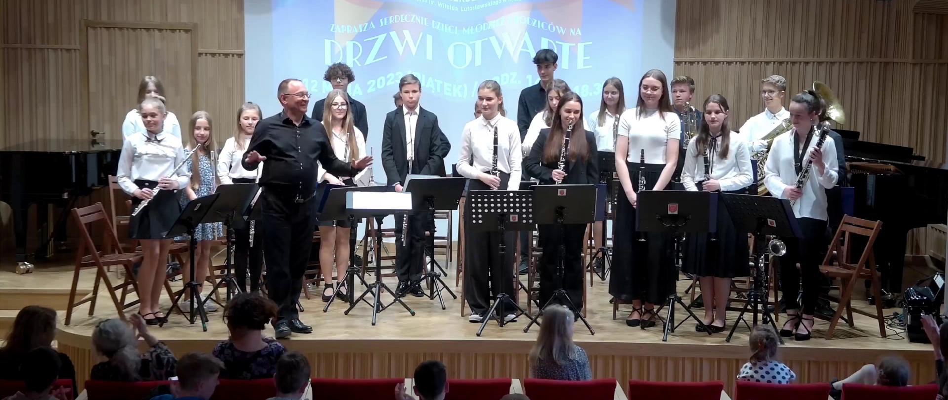Grafika - zdjęcie. Na estradzie sali koncertowej, przed siedzącą publicznością, stoją członkowie orkiestry dętej ze swoim dyrygentem. W tle wyświetlony jest baner "DRZWI OTWARTE".