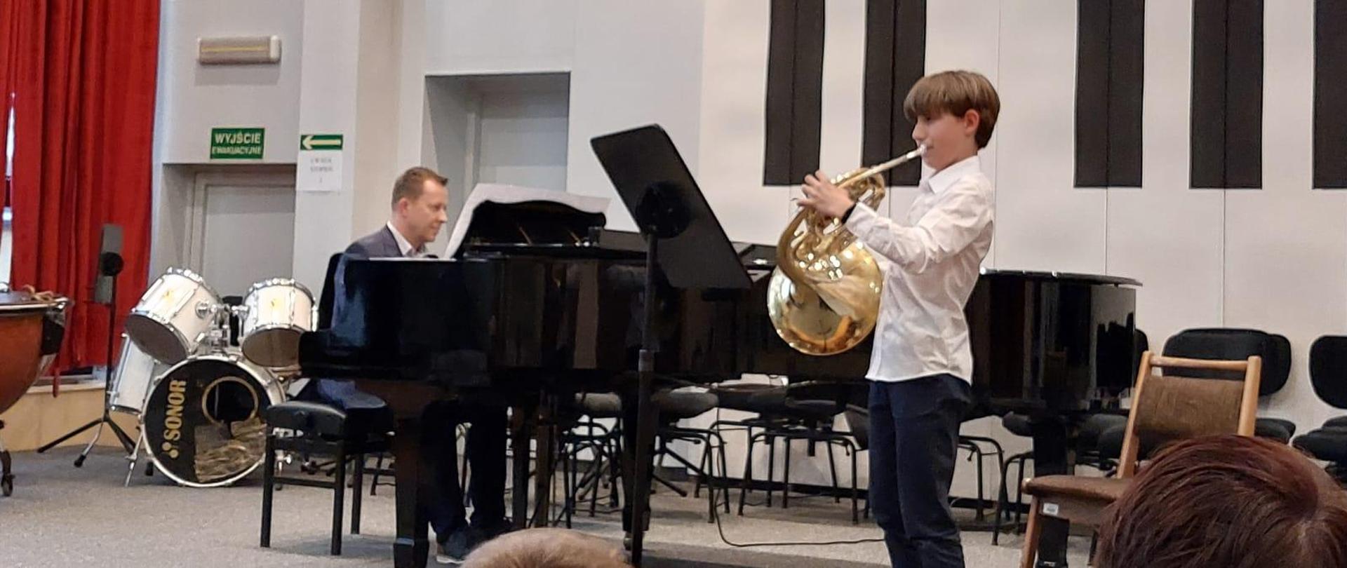 Chłopiec gra na waltorni, za nim na fortepianie gra mężczyzna.