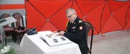 Komendant wojewódzki, generał, dokonuje wpisu w księdze pamiątkowej jednostki OSP. Na stole kronika, czapka, w tle parawan oddzielający wyposażenie remizy od miejsca uroczystości.