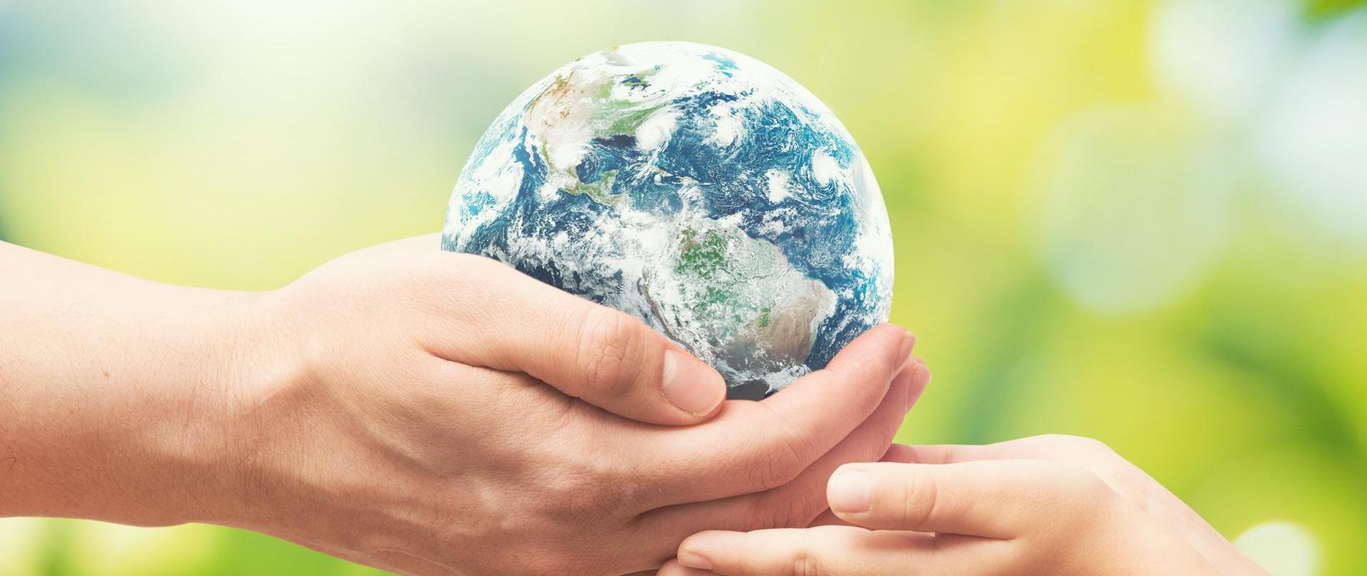 Na zdjęciu widoczne są dłonie osoby dorosłej oraz dziecka. W dłoniach osoby dorosłej znajduje się planeta Ziemia, która jest przekazywana do rąk osoby młodej. W tle widoczna jest roślinność. 