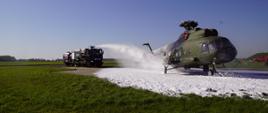 Zdjęcie przedstawia samochód Wojskowej Straży Pożarnej który z działka wodno-pianowego gasi pianą śmigłowiec.