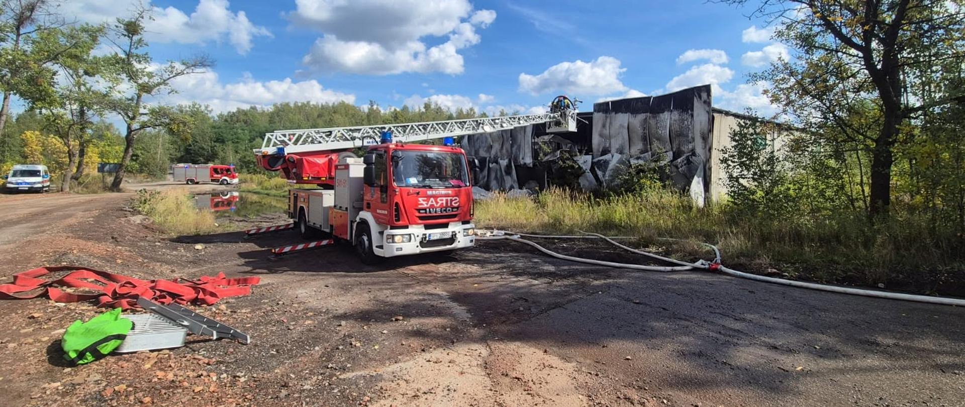 Na zdjęciu widoczny jest samochód pożarniczy wyposażony w drabinę mechaniczną, ustawiony przed spalonym budynkiem przemysłowym. Strażacy prowadzą działania gaśnicze z wykorzystaniem drabiny oraz linii wężowych. W tle znajdują się kolejne pojazdy ratowniczo-gaśnicze, a na pierwszym planie widoczny jest sprzęt wykorzystywany podczas działań.
