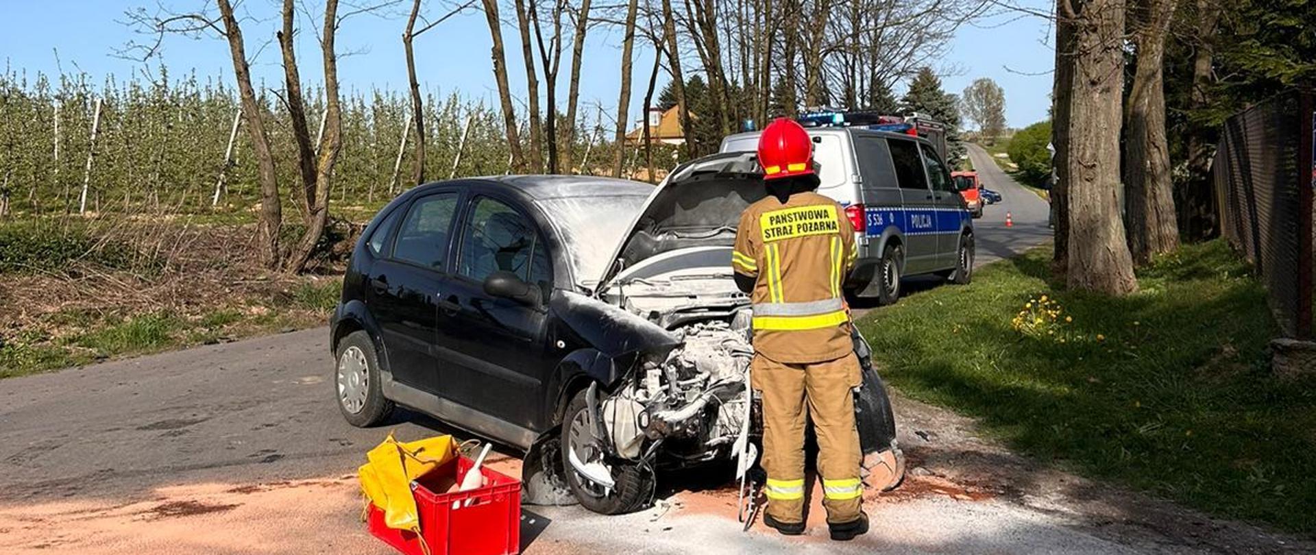 Na zdjęciu widoczny rozbity samochód i strażaka zabezpieczającego zdarzenia. Na drugim planie radiowóz policji