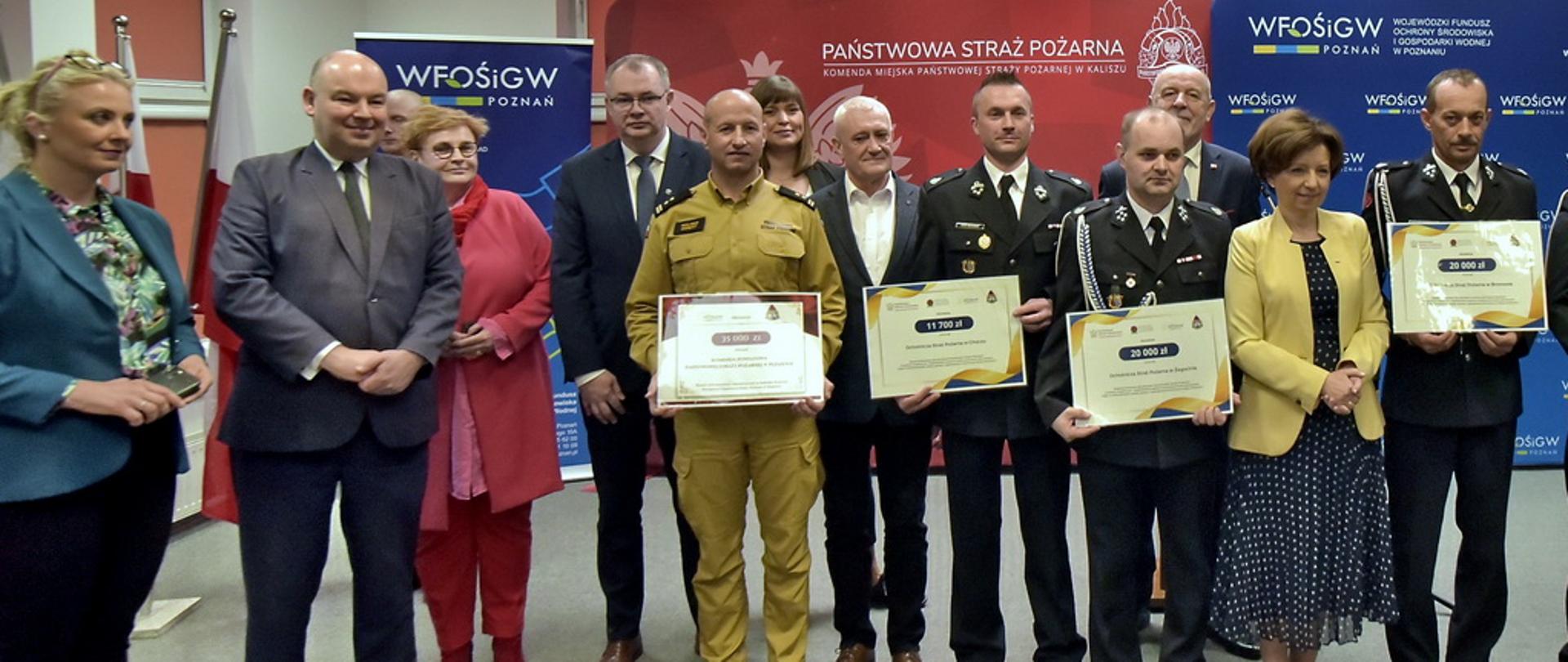 Na tle banerów z napisami państwowa straż pożarna i wojewódzki fundusz ochrony środowiska i gospodarki wodnej stoi w szeregu grupa mężczyn część z nich ubrana w mundury straży pożarnej trzyma w ręku promesy