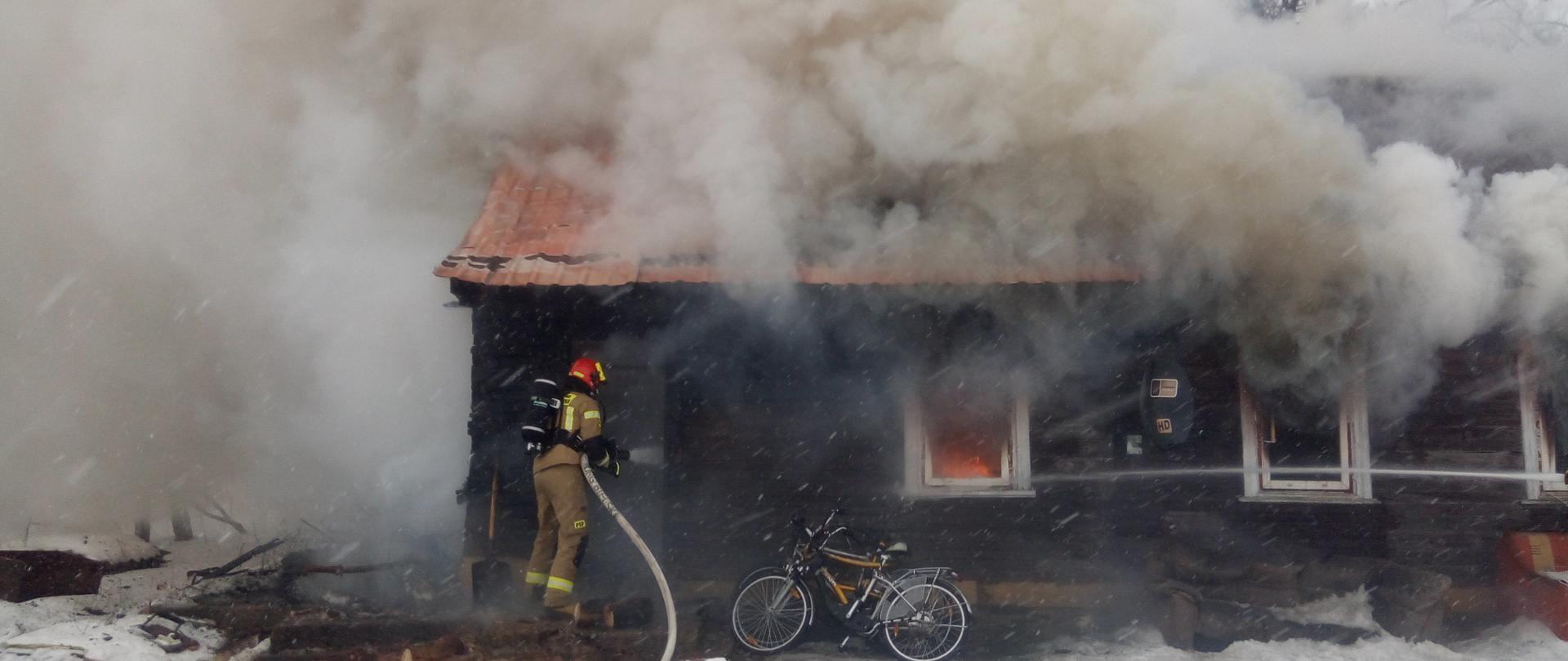 Drewniany budynek mieszkalny objęty pożarem. Przez okna wydobywa się gęsty dym, W jednym z okien widoczne płomienie. Górna część budynku zasłonięta dymem. Przed drzwiami strażak w ubraniu specjalnym koloru piaskowego, czerwonym hełmie i butlą powietrzną na plecach trzymają wąż strażacki prowadzi działania gaśnicze.