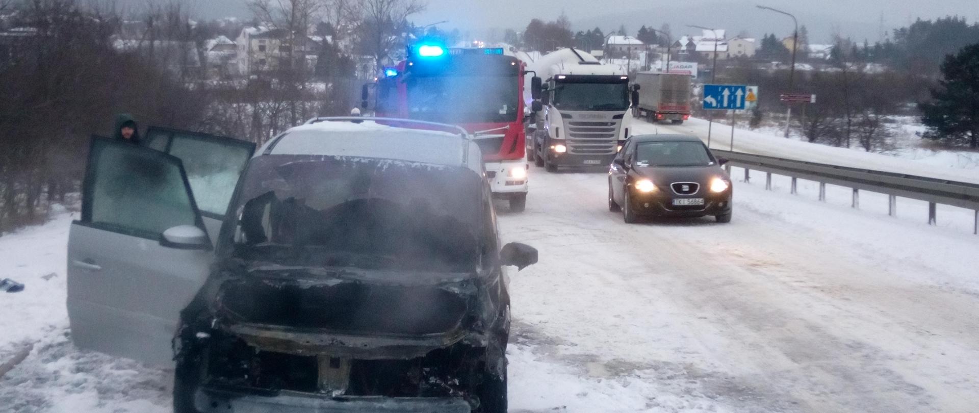 Na zdjęciu znajduje się spalony samochód marki Renault. Ma otwarte prawe drzwi - z jego wnętrza wydobywa się jeszcze dym. W tle samochód straży pożarnej na sygnałach. Jezdnia jest cała w śniegu.