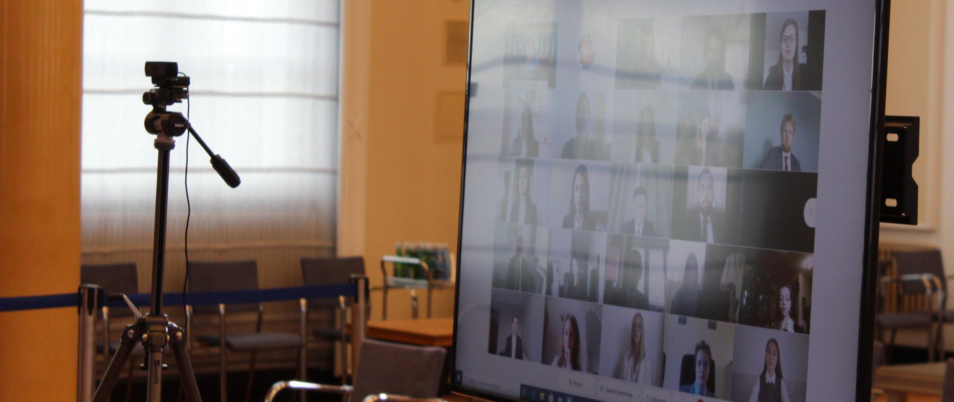 Ekran telewizora na którym wyświetlani są uczestnicy wideokonferencji. Obok kamera. W tle okno