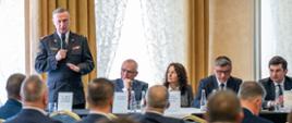 Na zdjęciu widać konferencję w sali. Na pierwszym planie znajduje się publiczność, głównie mężczyźni ubrani w garnitury, siedzący na krzesłach ustawionych w rzędach. Niektórzy uczestnicy są zwróceni w stronę prelegenta, inni pochylają głowy lub notują.
W tle, przy długim stole, siedzą paneliści – kobieta i kilku mężczyzn w eleganckich strojach. Każdy z nich ma przed sobą tabliczkę z imieniem i nazwiskiem. stoi i przemawia do mikrofonu, wykonując gest ręką. Za stołem stoi i przemawia do mikrofonu, ubrany w mundur wyjściowy st. bryg. Damian Legierski - pełniący obowiązki śląskiego komendanta wojewódzkiego PSP.
Za panelistami znajdują się duże okna przysłonięte ozdobnymi, jasnymi zasłonami, przez które wpada naturalne światło.
