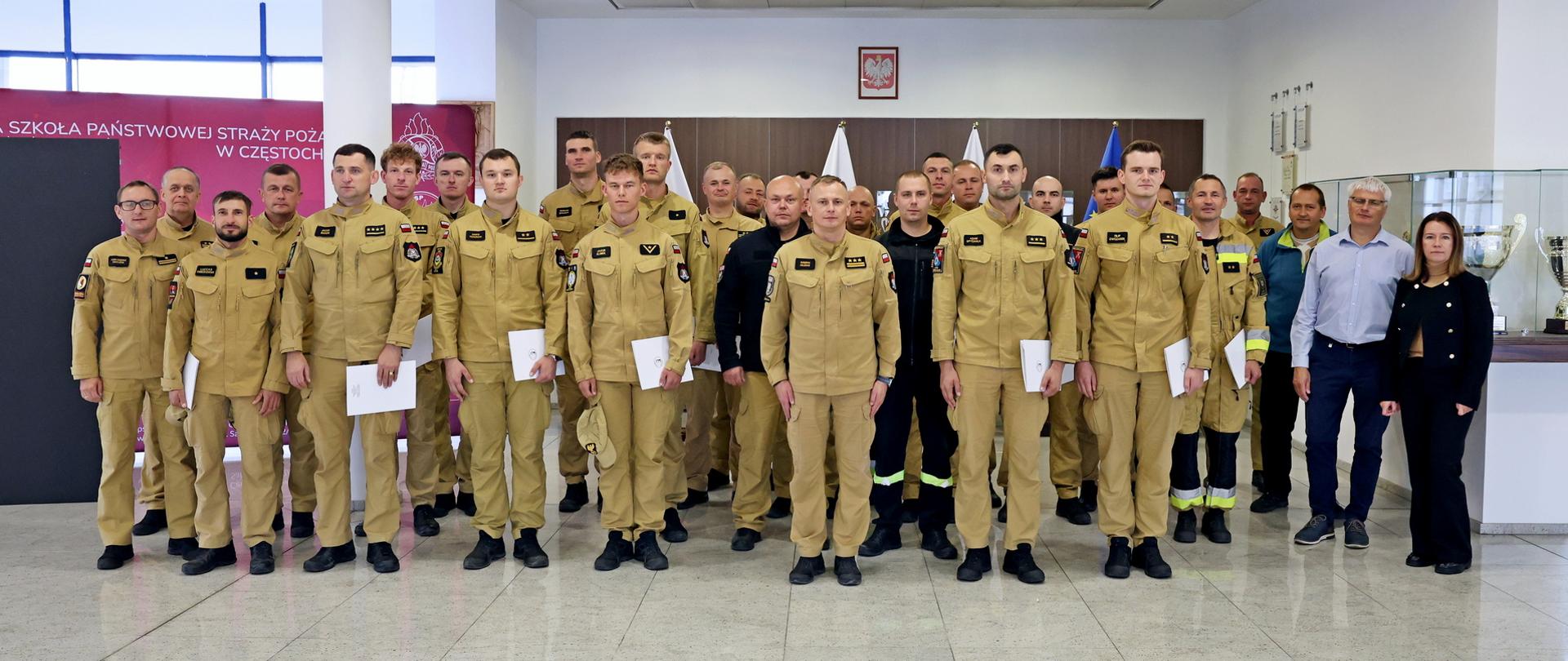 Pamiątkowe zdjęcie grupowe funkcjonariuszy PSP będących uczestnikami szkolenia z zakresu awaryjnego rozładunku cystern w towarzystwie Zastępcy Komendanta Centralnej Szkoły PSP, kobiety, instruktorów szkolenia oraz dwóch funkcjonariuszy CS PSP