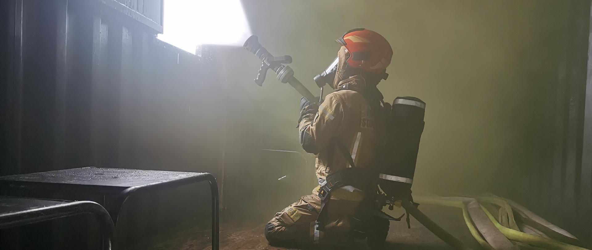 strażak w mundurze bojowym, masce ochrony dróg oddechowych w hełmie klęczy i trzyma wąz z lejącą się wodą, strażaka znajduje się w pomieszczeniu