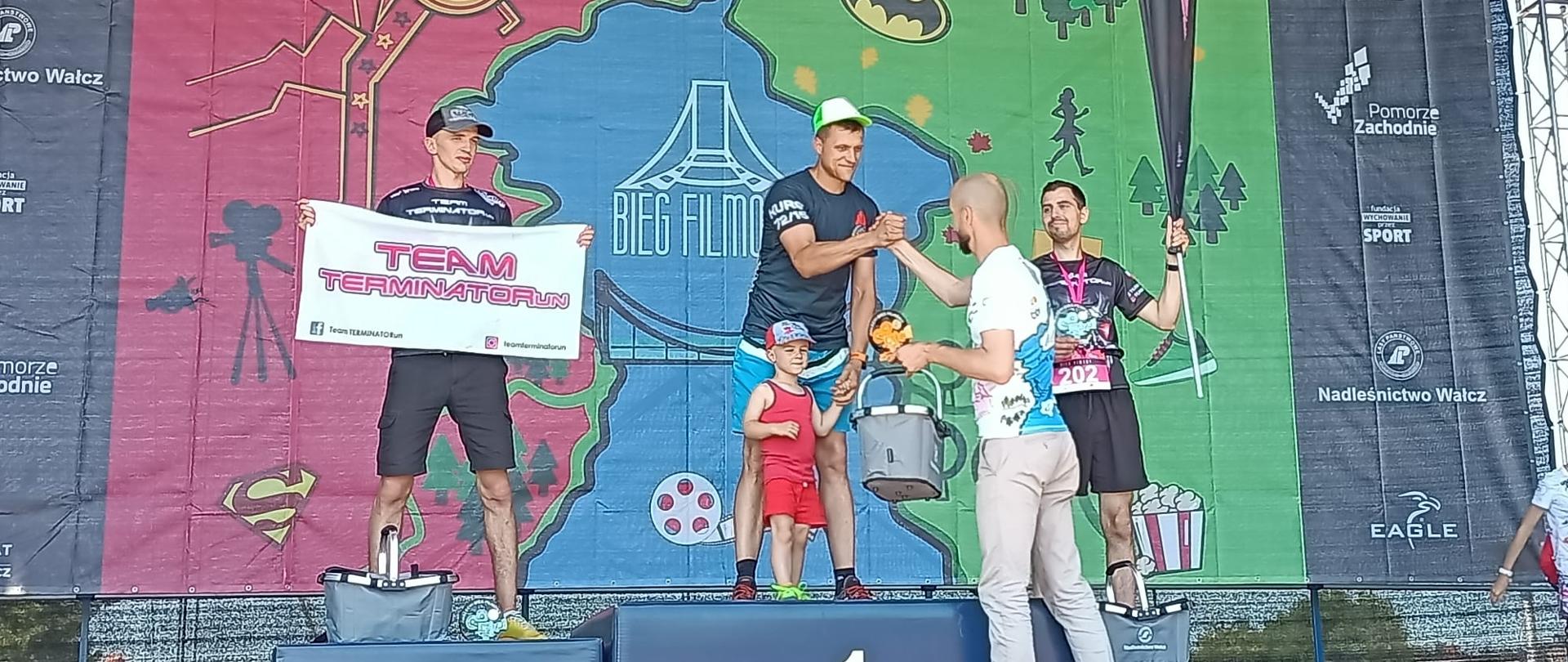 Zdjęcie przedstawia zwycięzców biegu, na najwyższym podium ogn. Maciej Malec z KP PSP w Wałczu oraz wręczenie nagrody na scenie Wałeckiego Festiwalu Biegowego 