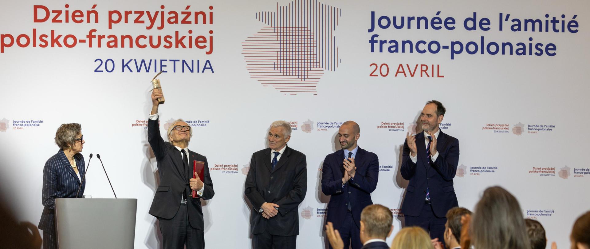 Andrzej Seweryn in a suit raises a statuette during the Polish-French Friendship Day ceremony, standing on stage next to other participants who are applauding. On the stage French Minister Delegate for Europe Benjamin Haddad and Secretary of State at the Polish Ministry of Foreign Affairs Ignacy Niemczycki