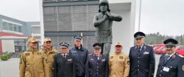 Grupa strażaków z Polski i Niemiec stoi na tle pomnika żołnierza,w dalszym planie budynek i czerwone samochody.