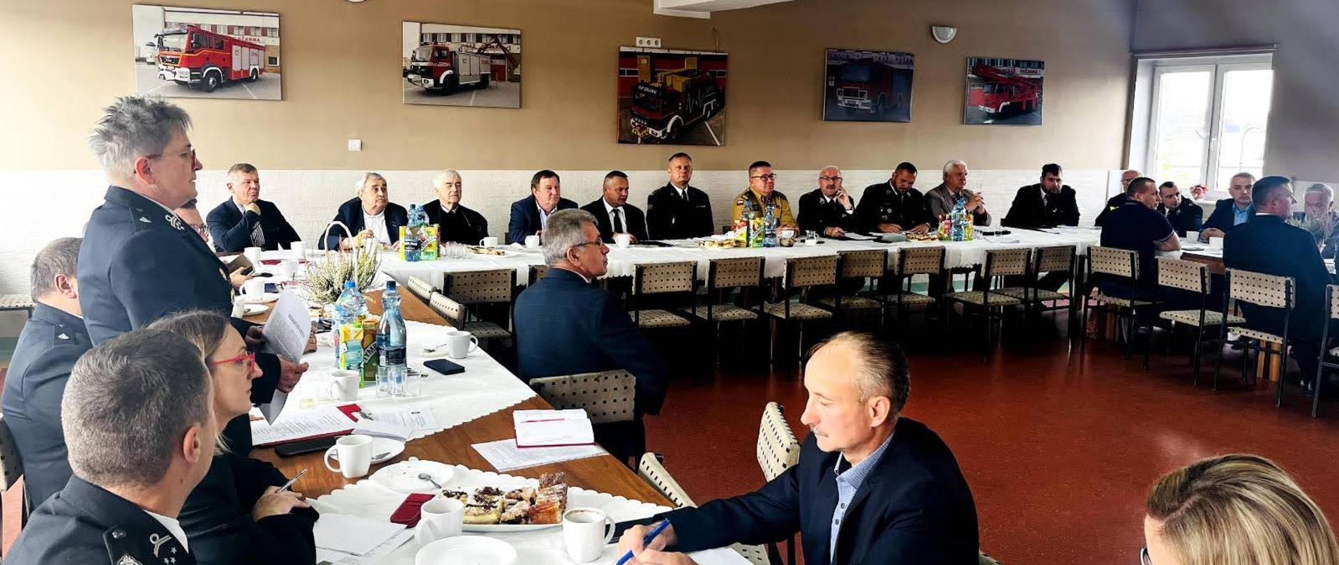 Druhowie ochotniczych straży pożarnych siedzą za stołami podczas spotkania w spotkaniu uczestniczą funkcjonariusze Państwowej Straży Pożarnej.