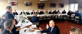 Druhowie ochotniczych straży pożarnych siedzą za stołami podczas spotkania w spotkaniu uczestniczą funkcjonariusze Państwowej Straży Pożarnej.