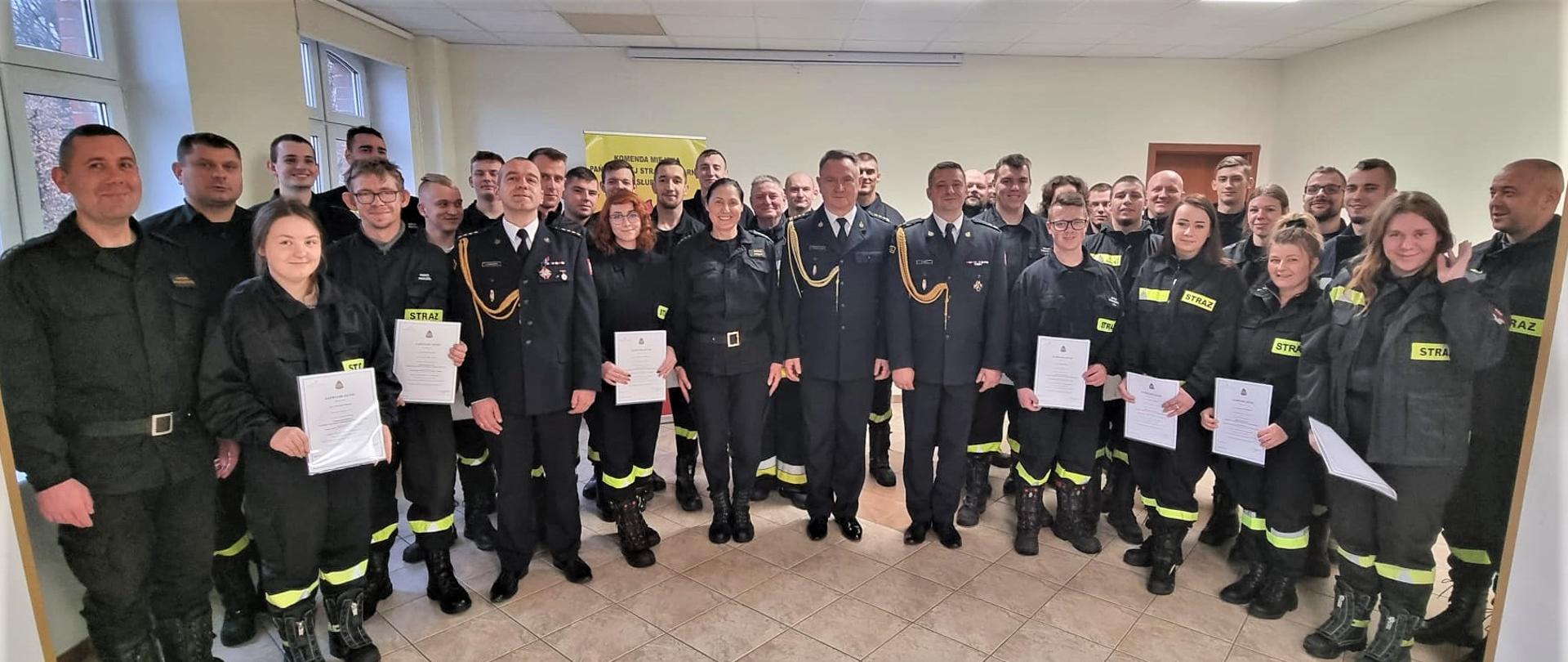 Zdjęcie przedstawia wszystkich uczestników szkolenia podstawowego strażaka ratownika Ochotniczych Straży Pożarnych wraz z st. bryg. Tomaszem Siemiętowskim, Zastępcą Pomorskiego Komendanta Wojewódzkiego PSP, mł. bryg. Krzysztofem Trockim, Komendantem Miejskim PSP w Słupsku oraz st. kpt. Iwoną Lenart, kierownikiem szkolenia. Absolwenci kursu trzymają w rękach stosowne zaświadczenia.