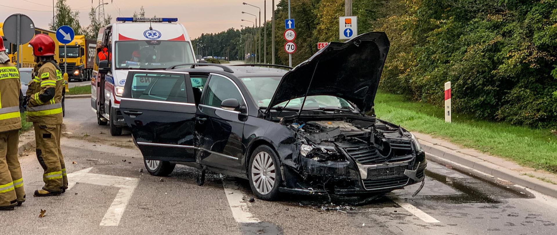 Zdjęcie przedstawia pojazd uszkodzony w wyniku wypadku drogowego, w tle karetka pogotowia oraz strażacy.