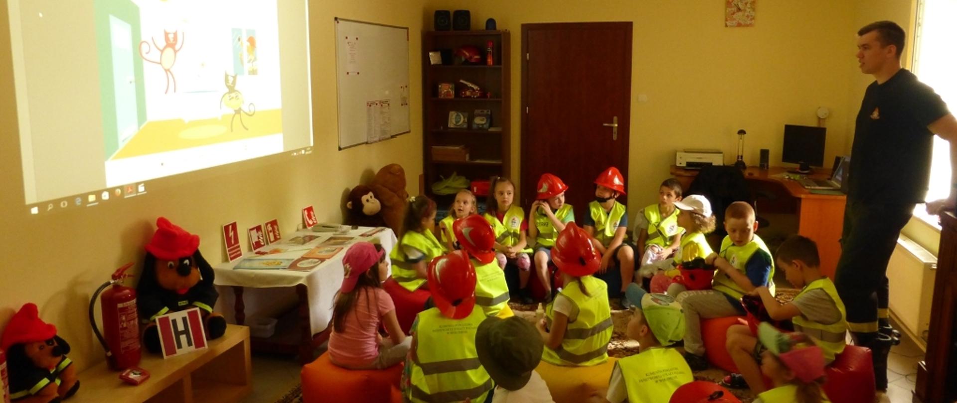 Na zdjęciu prowadzący zajęcia dla dzieci strażak z KP PSP Wołomin oraz grupa dzieci podczas oglądania prezentacji na ścianie sali. Dzieci ubrane w kamizelki odblaskowe i hełmy strażackie siedzą na pufach. 