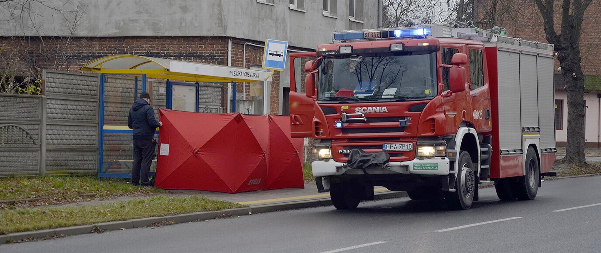 Na zdjęciu wóz strażacki, obok niego przy przystanku autobusowym rozstawiony przez strażaków czerwony parawan. Za nim strażacy udzielają pomocy osobie poszkodowanej.