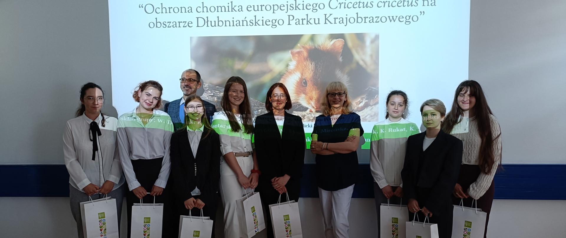 Grupa osób, kobiet i mężczyzn pozuje do zdjęcia na tle ściany na której wyświetlany jest obraz chomika europejskiego. Widać również napis: "Ochrona chomika europejskiego Cricetus cricetus na obszarze Dłubniańskiego Parku Krajobrazowego."