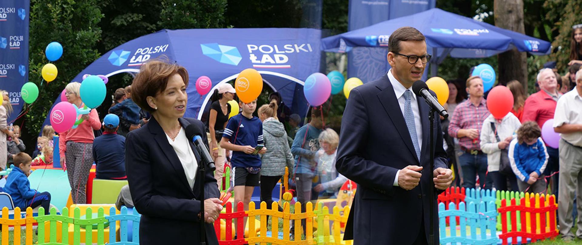 Na zdjęciu premier Morawiecki i minister Maląg. w tle bawiące się dzieci. Widać kolorowe balony w rękach, kolorowy płotek oddzielający miejsce zabawy dzieci. 